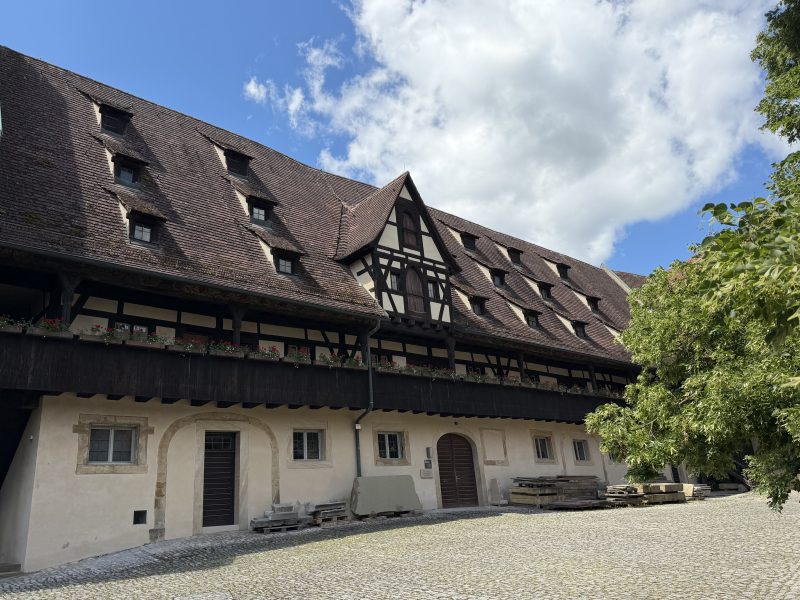 Historisches Fachwerkgebäude an einem gepflasterten Hof unter blauem Himmel.
