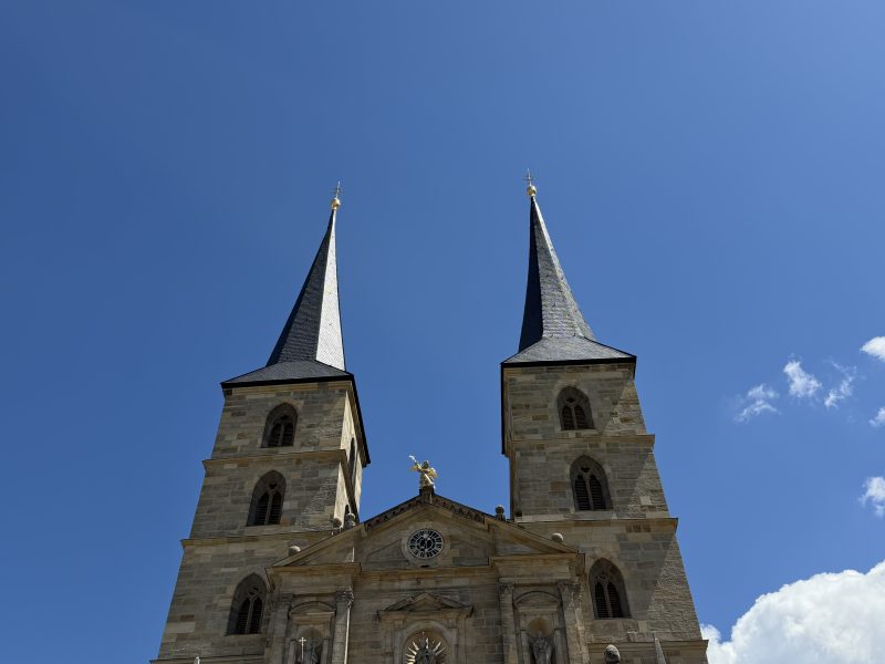 Zwei hohe Kirchtürme und eine steinerne Kirchenfassade vor blauem Himmel mit wenigen Wolken.
