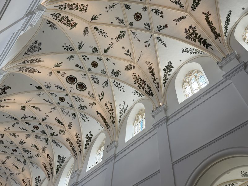 Helles Kirchengewölbe mit floralen Verzierungen und hohen Rundbogenfenstern.