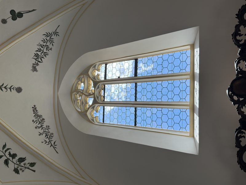 Hohes Fenster mit blauem Himmel dahinter, umgeben von hellen Wänden und feinen Deckenverzierungen.