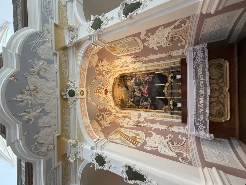Reich verzierter Altarraum in einer Kirche mit Stuck, Gemälden und hellen Wandflächen.